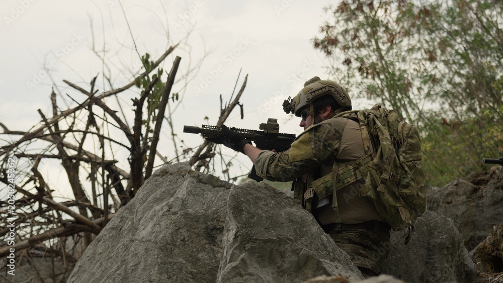 Fototapeta premium A special forces soldier in full tactical gear aims an assault rifle from behind a rock, conducting surveillance during a covert military operation outdoors.