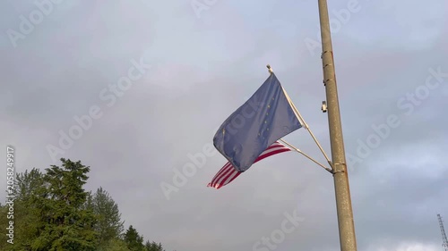 The flag of the state of Alaska with the United States flag behind, waving gently from a pole. 