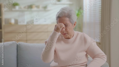 Asian senior woman having experiencing an intense headache or migraine and pressing temples, sitting on couch at home feeling unwell. appearing to have headache or discomfort.  health insurance.