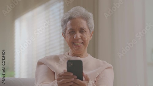 elderly Asian woman relaxing on sofa with phone in living room. Connect internet at home Wi-Fi, play social media. transfer money online or financial payment, smile or laugh with happy and positive.