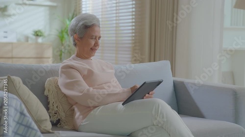 elderly Asian woman relaxing on sofa with tablet in living room. Connect internet at home Wi-Fi, play social media. transfer money online or financial payment, smile or laugh with happy and positive.