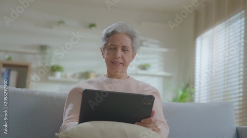 elderly Asian woman relaxing on sofa with tablet in living room. Connect internet at home Wi-Fi, play social media. transfer money online or financial payment, smile or laugh with happy and positive.