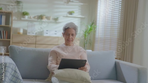 elderly Asian woman relaxing on sofa with tablet in living room. Connect internet at home Wi-Fi, play social media. transfer money online or financial payment, smile or laugh with happy and positive.