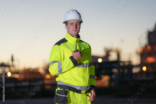 Industrial worker safety helmet high visibility jacket radio refinery at dusk, male engineer holding walkie talkie, reflective protective clothing, industrial plant background