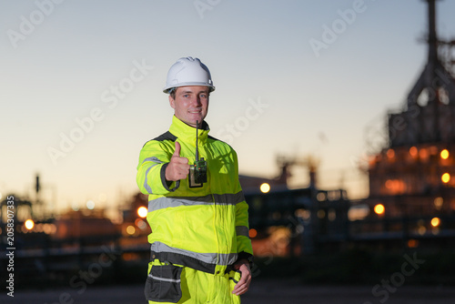 Industrial worker safety hard hat high visibility thumbs up refinery dusk male engineer wearing protective clothing standing near industrial plant lights confident approval gesture
