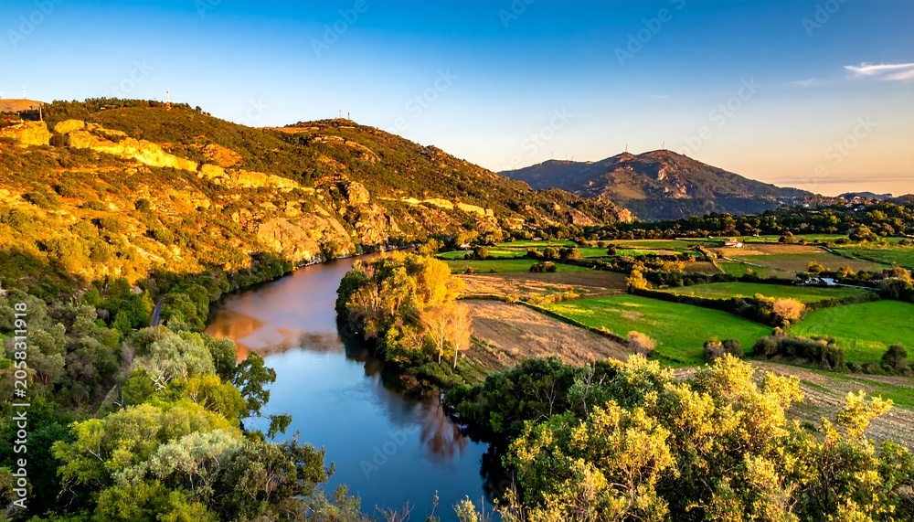 Obraz premium A serene river meanders through a picturesque valley surrounded by lush greenery and rolling hills under a clear blue sky