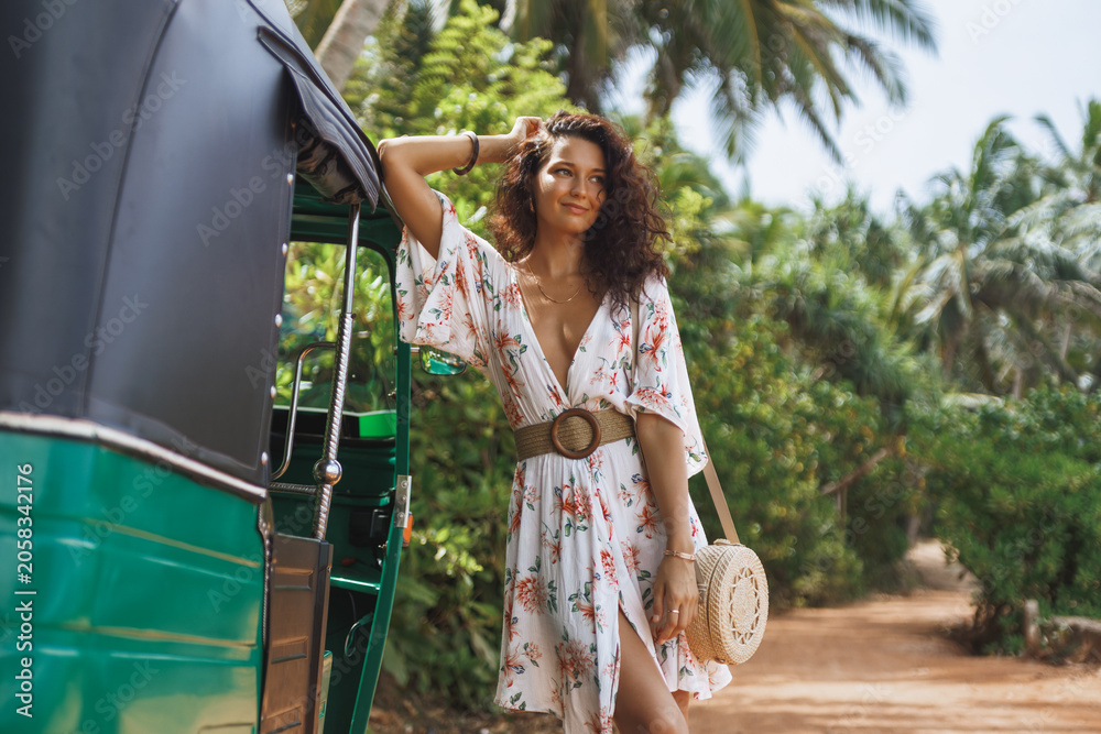 Fototapeta premium Woman in floral dress standing by green tuk-tuk on tropical dirt road