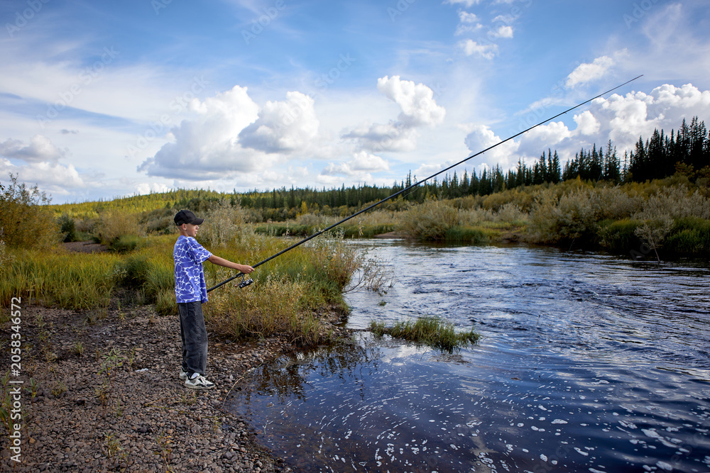 Naklejka premium A young boy stands on the rocky bank of a river while fishing with a long rod under a dramatic blue sky filled with white clouds. He enjoys a peaceful outdoor adventure in nature.