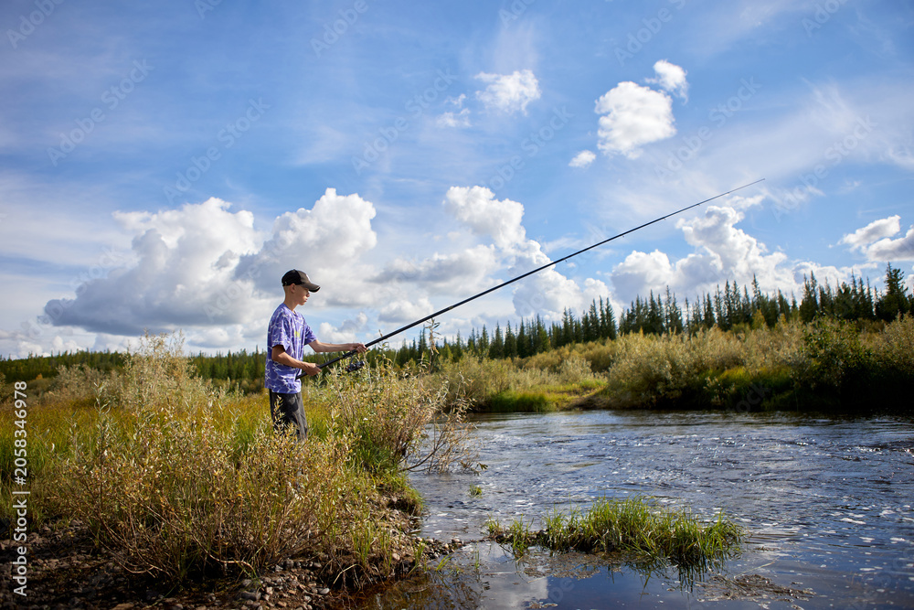 Naklejka premium A teenage boy fishing with a long rod on a scenic riverbank under a dramatic blue sky. Surrounded by wild grasses and forest, he enjoys a peaceful outdoor recreation moment in untouched nature.
