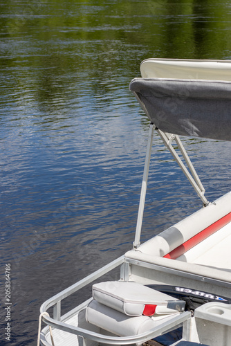 Close up view of the stern corner railing and folded bimini top canopy of a pontoon boat. Summer recreation lifestyle scene featuring white marine seats and aluminum support frames next to a river.
