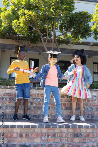 Three children wearing mortarboard caps holding red ribboned diplomas on brick wall at school