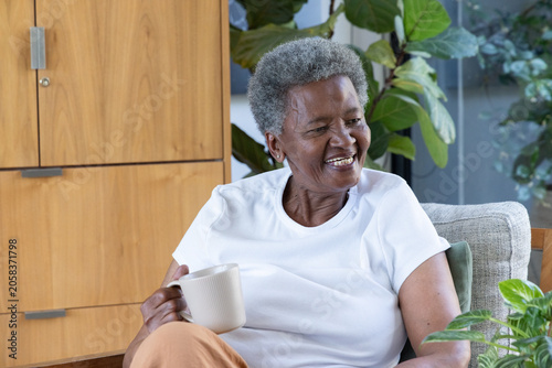 Senior African American woman sitting in armchair at home holding ribbed mug near potted plants