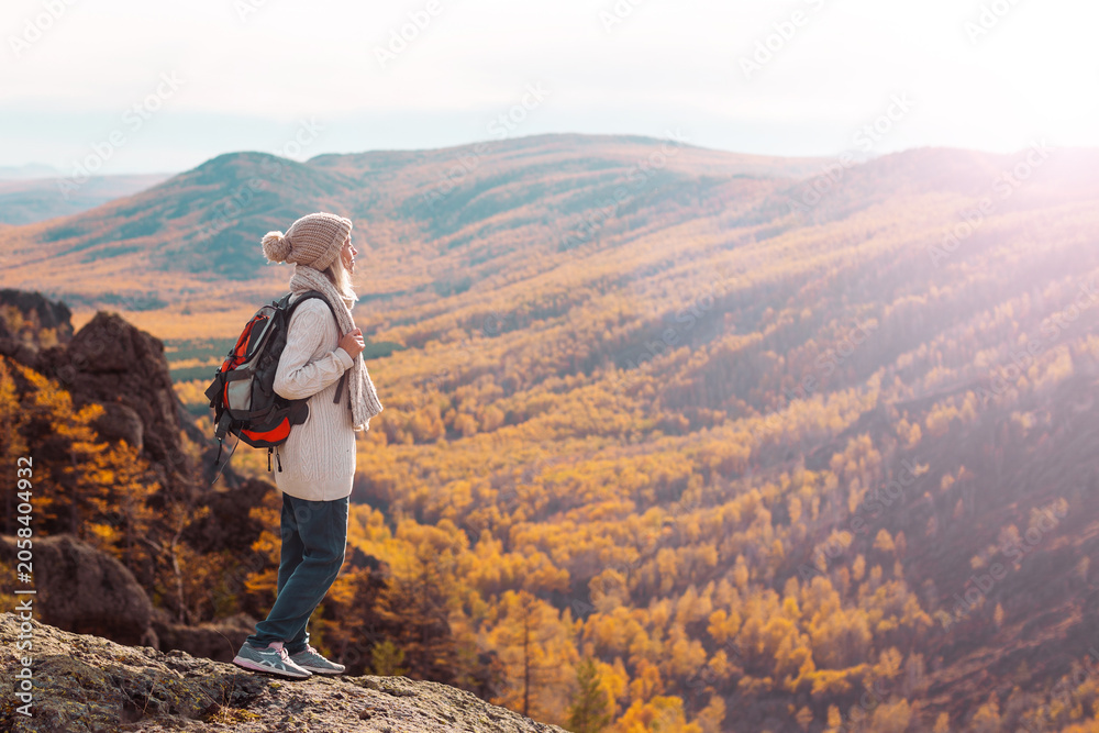 Naklejka premium Woman hiker in sweater and beanie standing on mountain cliff looking at autumn forest valley. Adventure traveler with backpack enjoying landscape scenic view. Active lifestyle travel concept.