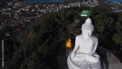 Massive white buddha statue overlooking town and harbor in drone angle, capturing relationship between monument and coastal community with sunlit roofs and winding roads creating vibrant context 