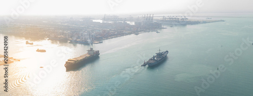 Fotografie naval warship escorting a large commercial container ship near a busy industrial port, symbolizing maritime security, naval patrol, global trade protection, and geopolitical stability