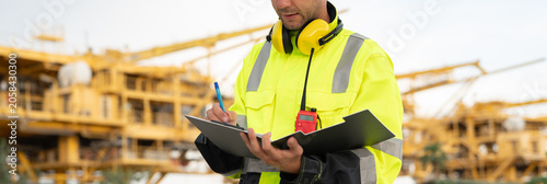 Industrial engineer holding a check list clipboard and walkie-talkie at an offshore energy platform. Concept of workplace safety compliance, power infrastructure, and professional engineering