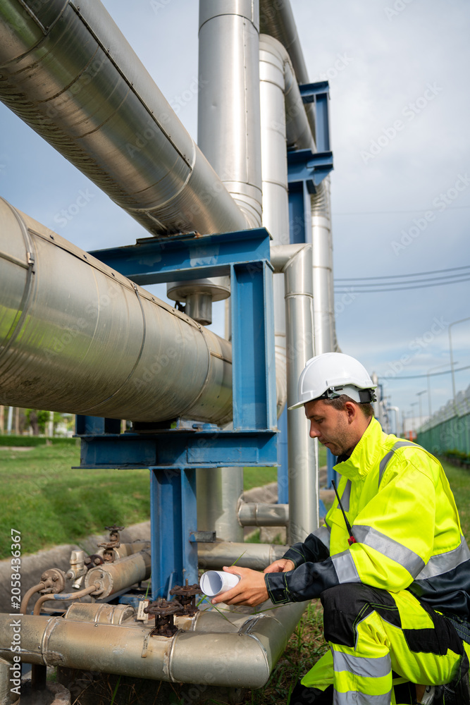 Naklejka premium industrial engineer wearing a white hard hat and high-visibility PPE squats to point at a large metal pipeline while speaking into a walkie-talkie during an outdoor refinery inspection.