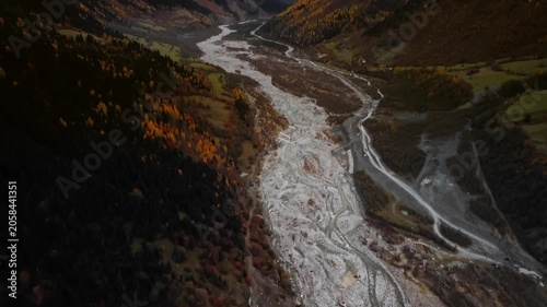 Aerial autumn braided river cutting valley in Svaneti Georgia, winding gravel channels, steep granite banks, golden larch forests, winding mountain road, textured sediment patterns, late afternoon