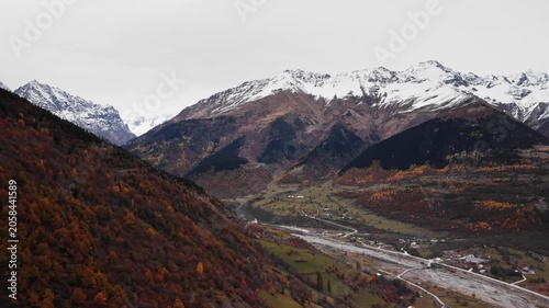 river valley flanked by autumn slopes and snow lined peaks with braided channel cutting through rocky basin, drone vantage highlights scale and geological textures, moody sky and crisp air convey