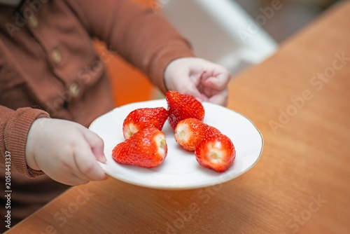 A child holding a white ceramic saucer filled with fresh, large ripe red strawberries