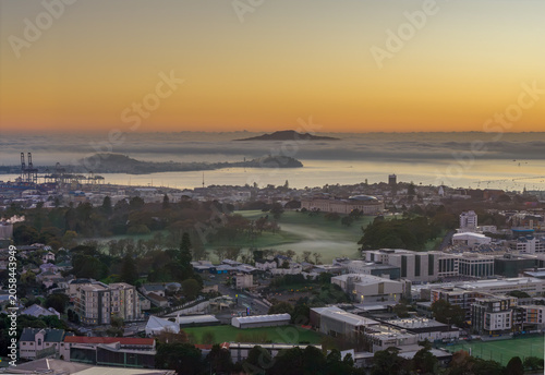 Auckland, New Zealand. Sunrise over the city skyline with fog rolling in from the harbor, casting a mystical glow on the port and surrounding buildings.