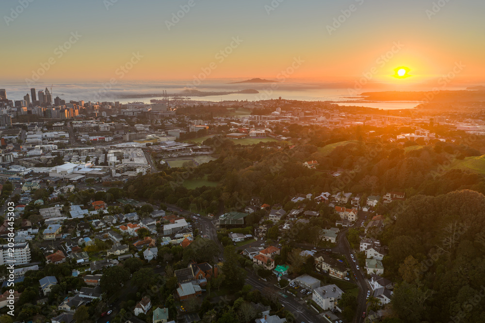 Fototapeta premium Sunrise over Auckland, New Zealand. The city skyline emerges from a layer of low clouds as the sun casts a warm glow over the harbor and surrounding hills.