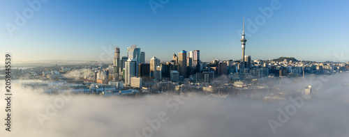 A stunning aerial view of Auckland, New Zealand, as low-lying fog blankets the harbor and city center, revealing the iconic Sky Tower and skyscrapers on a clear morning.