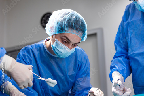 Young focused female surgeon in blue scrubs, surgical mask and cap holds forceps with gauze during surgery, assisted by surgical team in hospital OR.