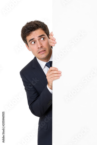 Anxious Young Businessman Peeking from Behind Blank Poster with Copy Space
