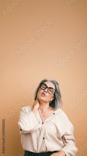 Mature stylish woman with grey hair and glasses massaging her sore neck. Exhausted lady feeling tired due to neck pain and muscle tension. Vertical