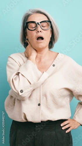 Middle aged business woman with gray hair stretching her neck, yawning from fatigue, and then smiling, isolated on a light blue background. Vertical