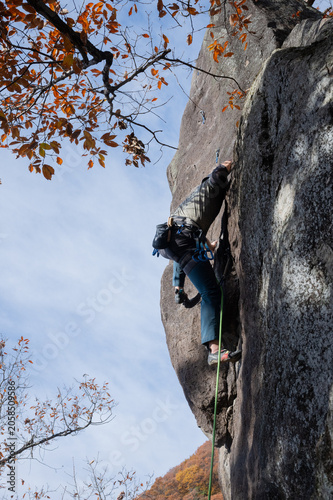 Rock climber ascending steep granite cliff face with autumn foliage and blue sky, low angle view
