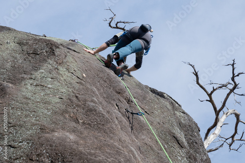 Rock climber scaling steep granite face with green rope and carabiners on sunny day