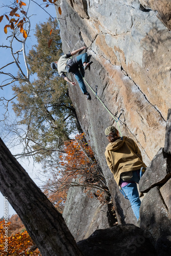 Rock climber scaling steep granite cliff face with belayer watching in autumn forest setting