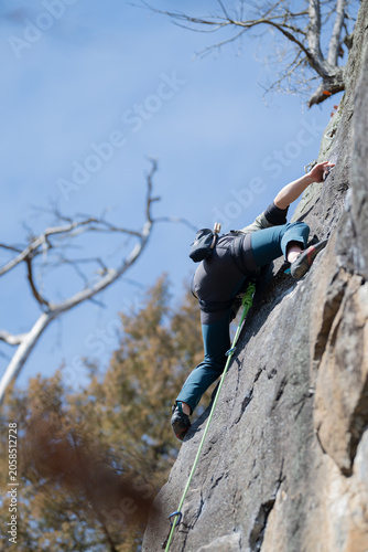 Rock climber scaling steep granite cliff face with green rope and harness surrounded by bare autumn trees against blue sky