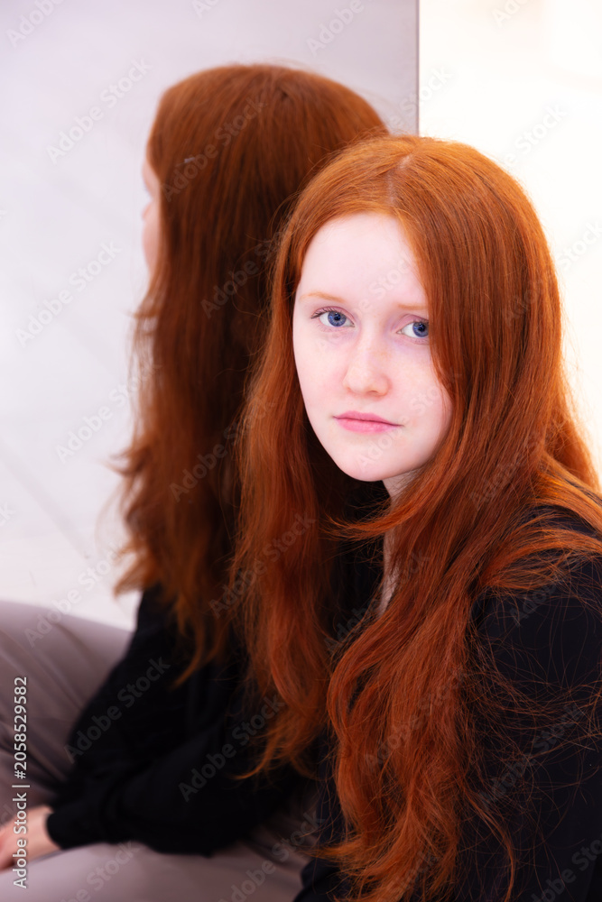 Naklejka premium Vertical portrait of pretty young blue-eyed girl with long wavy red hair and wearing black top and beige pants sitting next to her reflection in mirror 