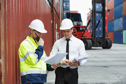 Two professionals assess shipping logistics within bustling container yard, reviewing documents and discussing operations while wearing hard hats and work attire for focused business setting.