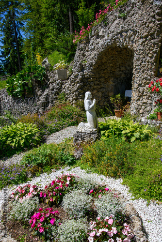 Obraz na plátně White stone statue of a praying nun in a peaceful garden setting with flowers and rustic stone wall, symbolizing faith, devotion, spirituality, prayer, and serenity