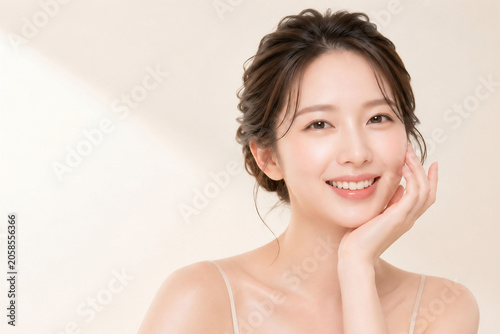やわらかな光に包まれた明るいベージュ背景でほほえむ若い女性の美容ポートレート。頬に手を添えたナチュラルメイクと透明感のある素肌を印象的に見せた、スキンケア・ビューティー・保湿イメージ