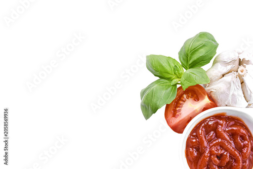 Tomato sauce in a white bowl isolated on white background. Red ketchup or marinara dip with fresh tomato slice, basil leaves and garlic clove, copy space.
