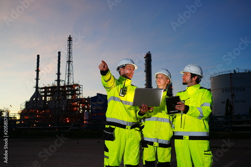 Obraz na plátně A team of two male and one female Caucasian engineer, wearing uniforms and helmets, stood working on laptops, inspecting a petrochemical refinery and working overtime