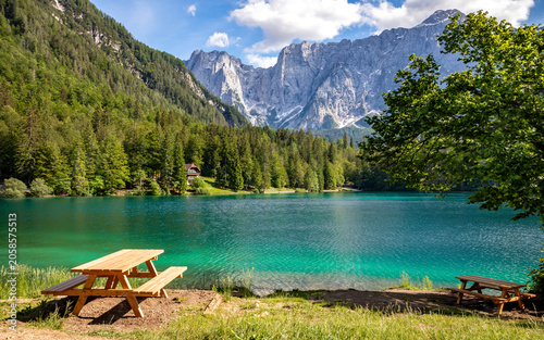 Turquoise alpine lake at Laghi di Fusine, along Sentiero del Lago Inferiore hiking trail, surrounded by forest and dramatic mountains with crystal-clear turquoise water. Idyllic alpine landscape.