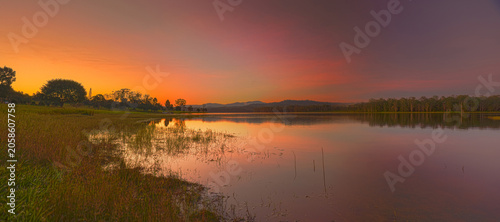 A wide panoramic sunset view over Lake Tinaroo in the Atherton Tablelands, Far North Queensland, with warm orange and purple evening light reflecting across calm water, distant hills, lakeside trees