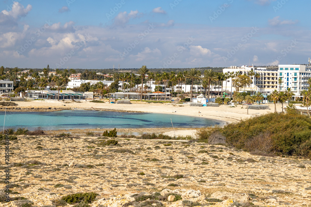 Fototapeta premium Turquoise water of Vathia Gonia beach lagoon during sunny winter morning. Empty sand coast with resort hotels and palm trees in low season, panoramic view, Ayia Napa, Cyprus.
