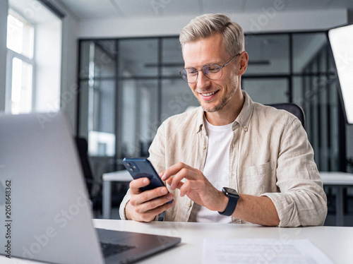Tableau sur toile Smiling blonde businessman uses his smartphone at a modern office desk, reflecting digital work engagement