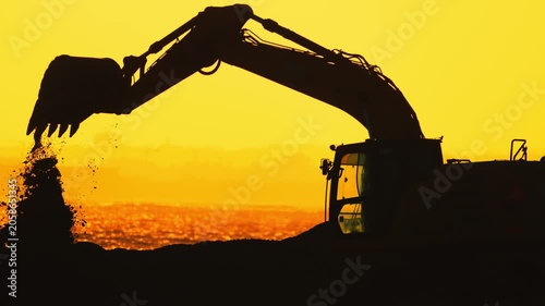 Excavator silhouette lifting earth against a vibrant sunset, showcasing the continuous motion of the arm and bucket in a construction site setting by the water