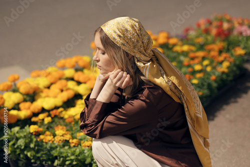 Schilderij op canvas Stylish blonde woman in a brown silk shirt and yellow babushka headscarf squatting by marigold flower beds at an outdoor street market