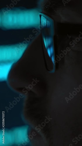Side view close up, focused software engineer in eyeglasses looking at blue code running on screen in dark room, programming script reflects in his lenses. AI and data science concept. Vertical video