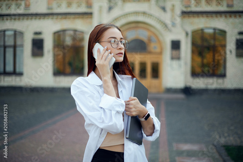 Businesswoman talking on phone outside a building while holding a notebook and wearing a white shirt