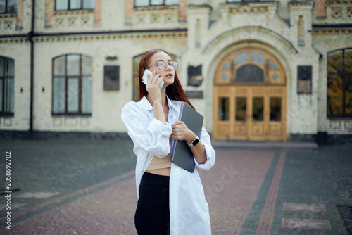 Businesswoman talking on phone outside a building while holding a notebook and wearing a white shirt